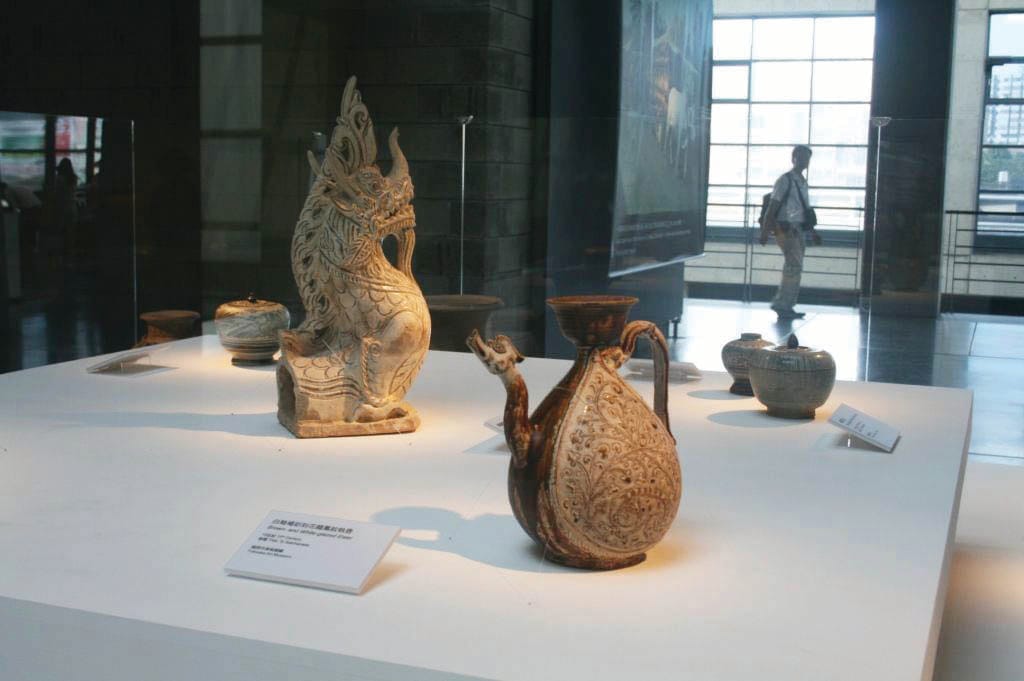 Ceramic artifacts are displayed on a white pedestal in a museum; a person is seen walking in the background.